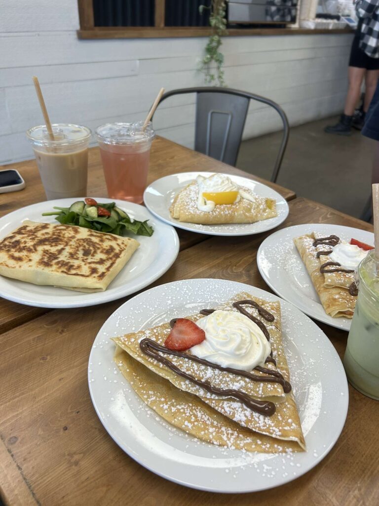 Angled photo of 4 different crepes and  3 different drinks. An iced coffee, a pink Italian soda, and an iced matcha in the corner of the picture. All on a wooden table/