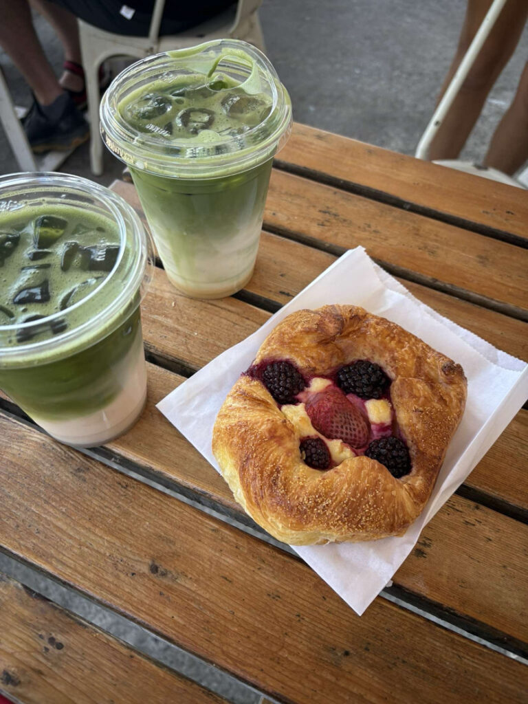 An angled birds eye view of 2 iced matchas in plastic to go cups and a danish with yellow custard and blackberries and strawberries in the middle laying on top of a napkin. All on a wooden table.