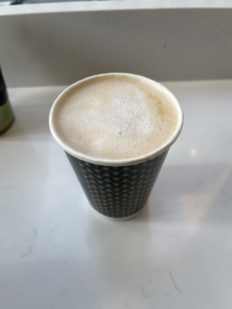 Image of a paper coffee cup that is green and has small gold sun patterns on it with foam on top of the cup. 
